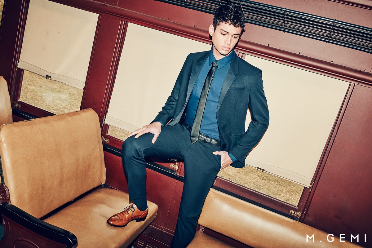 A young man poses in a blue suit with a black tie, seated on tan leather inside a train car near a window for a fashion editorial.
