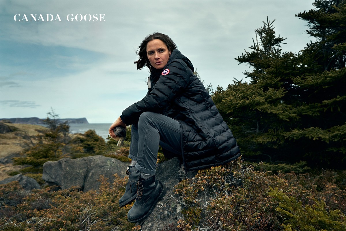 A woman in a black down jacket sits on rocks in an outdoor setting with trees, a body of water, and cloudy skies behind her.