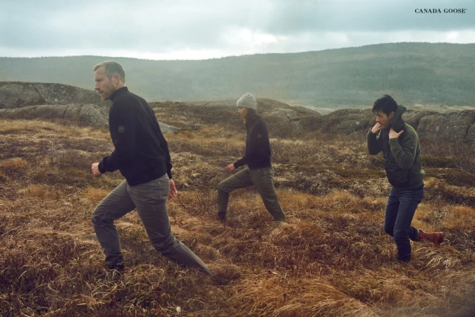 Three people wearing jackets and trousers walk through a field with a mountain range and cloudy sky in the background for Canada Goose.