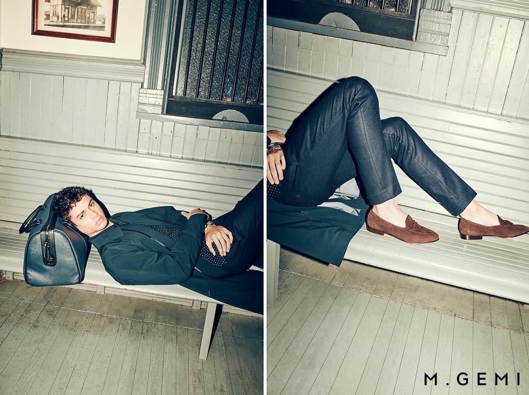 Male fashion model wearing a black suit and brown loafers, lying on a bench in an indoor station-like setting with natural lighting.