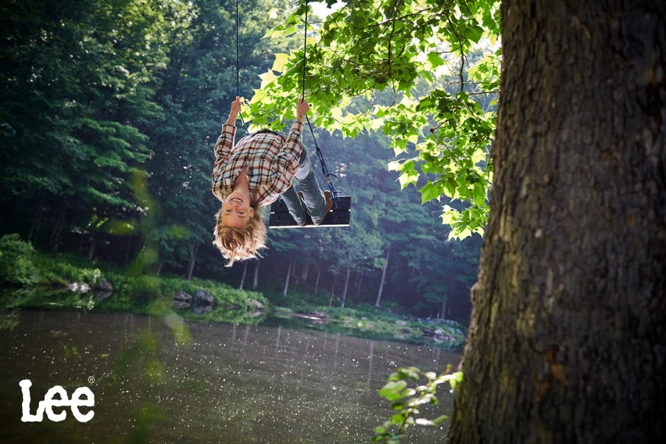 A cheerful child enjoys a tree swing, hanging upside down, in a forest with a serene lake. The light is bright and airy.