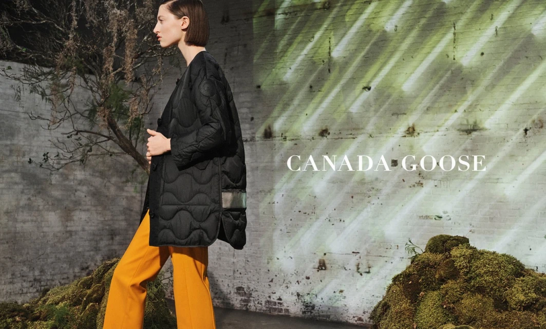 Woman models a Canada Goose black puffer jacket and orange pants in a studio with moss and a textured wall, showcasing fashion.