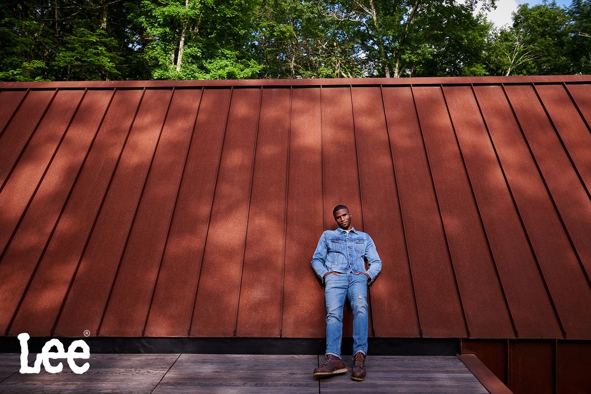 Man in denim jacket and jeans leaning against a corrugated rust-coloured metal wall, styled by Soraya Dayani.