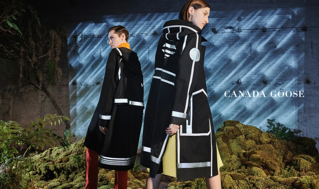 Two women model black and white Canada Goose coats in a mossy, naturalistic setting with a concrete wall and muted light.