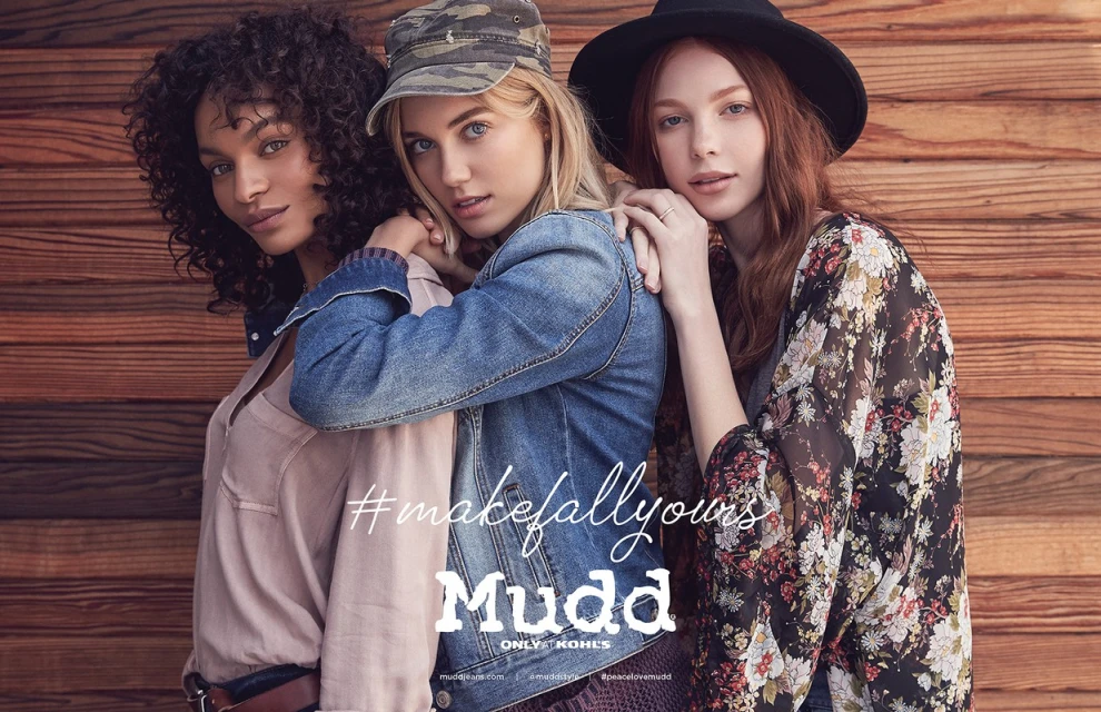 Three fashionable young women pose in a studio with a wood background. One wears a denim jacket, the others wear floral tops.