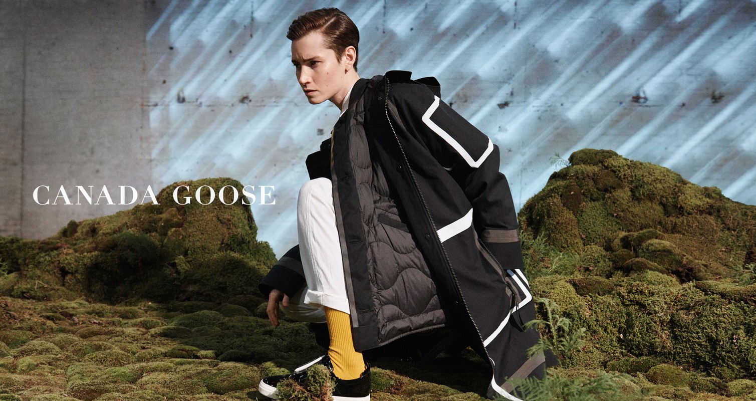 A young adult man models a black parka with white accents on a mossy hillside for Canada Goose fashion apparel.