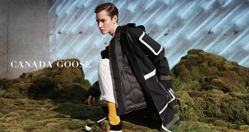 A young adult man models a black parka with white accents on a mossy hillside for Canada Goose fashion apparel.