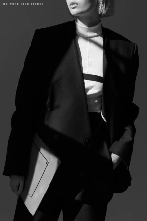Model in a black blazer and white turtleneck holding a light-colored folio against a dark studio background.