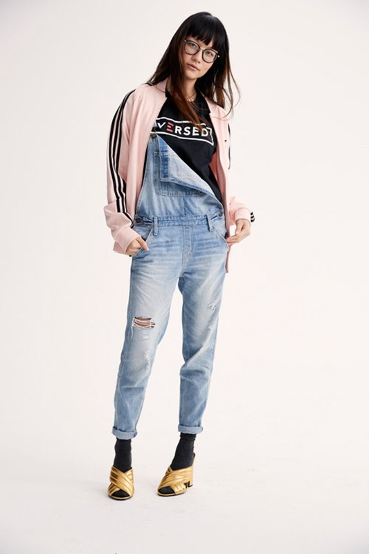 Full-length shot of a woman wearing a pink jacket, black top, denim overalls, and gold shoes, posing in front of a white backdrop.