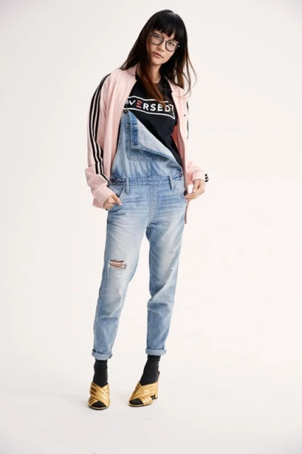 Full-length shot of a woman wearing a pink jacket, black top, denim overalls, and gold shoes, posing in front of a white backdrop.