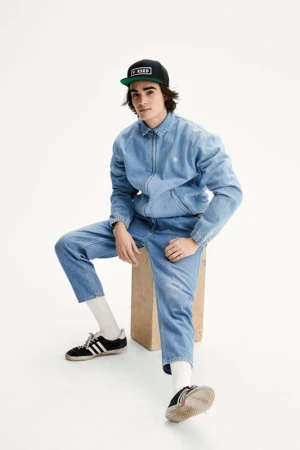 Young man in full denim outfit wearing a black baseball cap, seated on a wooden box against a white background.