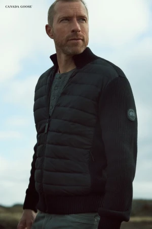 An adult male wears a black Canada Goose bomber jacket outdoors.  He is looking off camera in a medium shot with a blurred background.