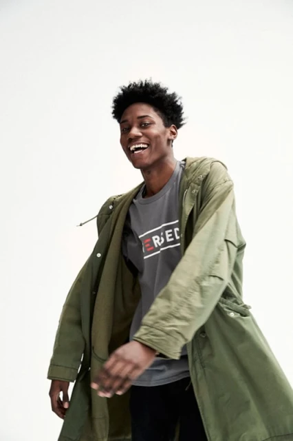 Young man smiles wearing olive green jacket and gray t-shirt in a bright studio. Casual fashion shoot against a white background.