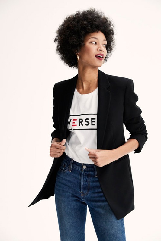 Woman with dark curly hair wearing a black blazer over a white graphic t-shirt and blue jeans