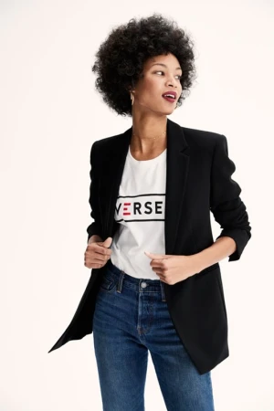 Woman with dark curly hair wearing a black blazer over a white graphic t-shirt and blue jeans