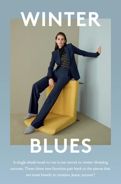 A woman in a blue suit poses on a yellow chair in a fashion editorial shot, wearing trousers and a striped sweater indoors.