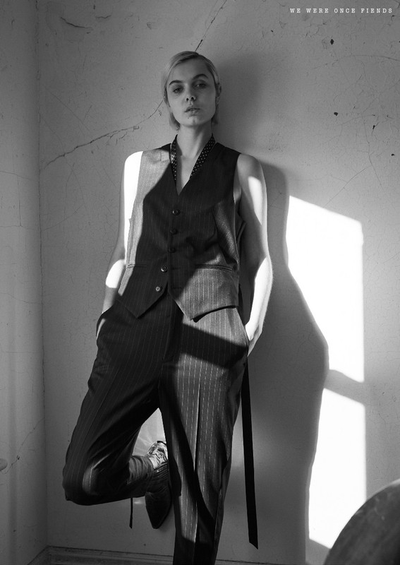 Black-and-white shot of a blonde woman leaning against a wall, wearing a vest suit and dress shoes, looking at the camera with a neutral expression.