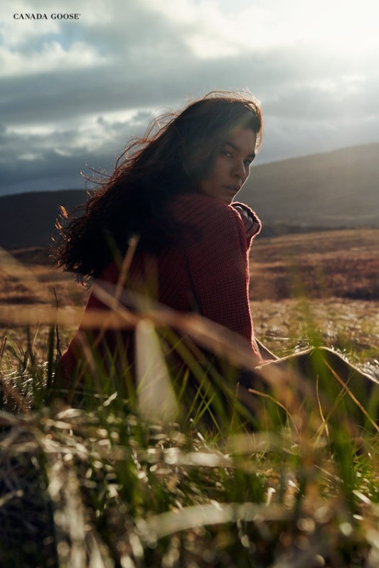 Brunette woman in a red jacket sits in a grassy field under a cloudy sky, illuminated by sunlight, in an outdoor fashion shot.