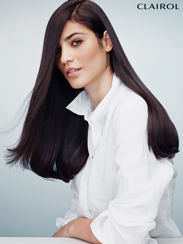 A woman with long, dark, straight hair poses in a white button-down shirt against a light background in a beauty advertisement.