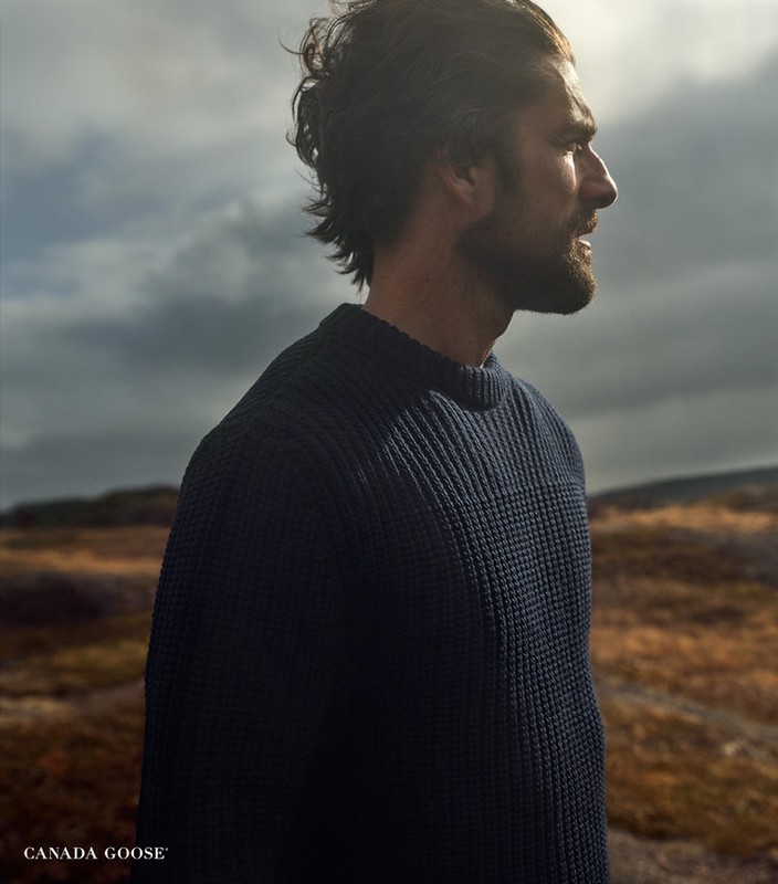 Profile shot of a dark-haired adult male wearing a navy sweater outdoors against a cloudy sky and field, promoting the Canada Goose brand.