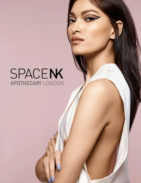 Headshot of a woman with dark hair and dramatic eyeliner against a lavender background, posing with arms crossed.