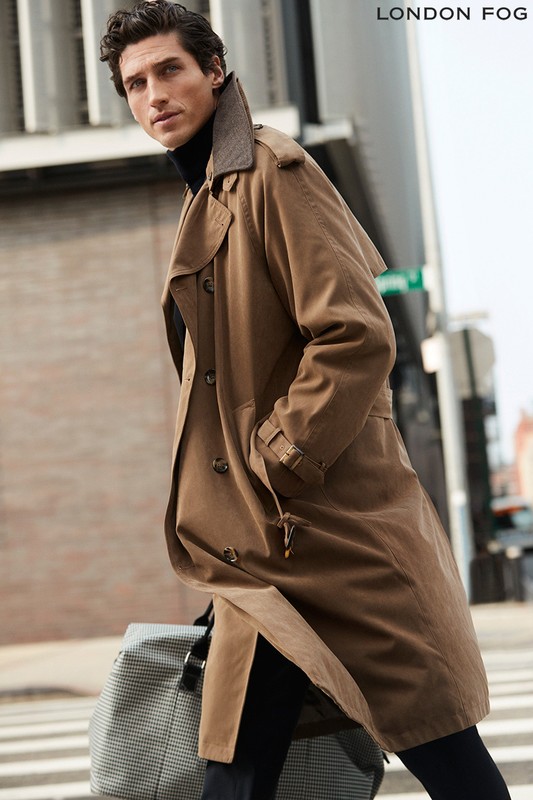 Man walking on a city street wearing a camel trench coat over a black turtleneck and carrying a patterned duffel bag