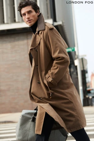 Man walking on a city street wearing a camel trench coat over a black turtleneck and carrying a patterned duffel bag