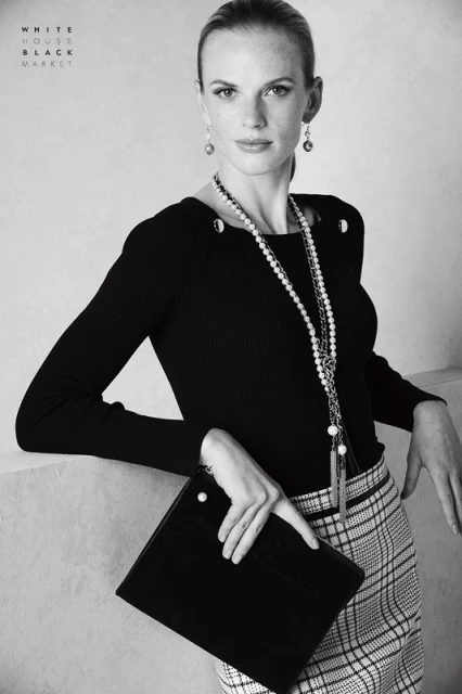 Elegant black and white portrait of a woman in a black top, plaid skirt, holding a black clutch, with a neutral background. 
