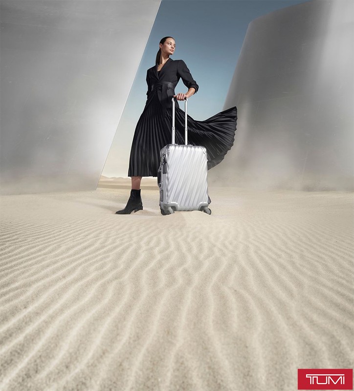 Fashion shot of a woman in a black dress pulling a silver Tumi suitcase in a desert landscape with geometric architectural structures.