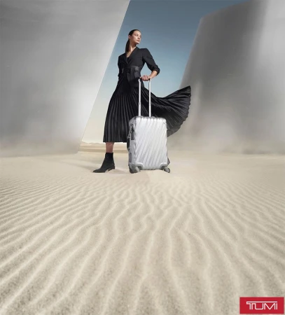 Fashion shot of a woman in a black dress pulling a silver Tumi suitcase in a desert landscape with geometric architectural structures.