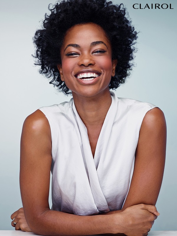 Smiling Black woman with afro wearing a sleeveless white wrap top with arms crossed in a beauty and fashion portrait against a light blue backdrop.