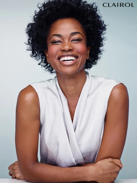 Smiling Black woman with afro wearing a sleeveless white wrap top with arms crossed in a beauty and fashion portrait against a light blue backdrop.
