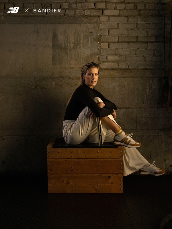 Woman in a black top and white pants sits on a wooden box, wearing New Balance sneakers, in front of a textured concrete wall.