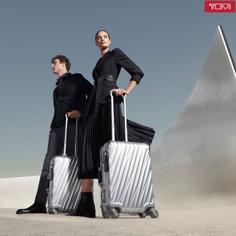 Two models present sleek silver suitcases in a minimalist desert setting under a clear sky, accentuating the luggage's modern design.
