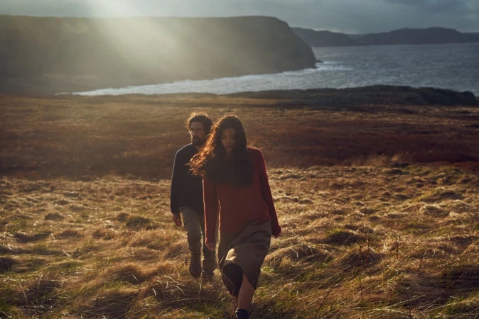 A man and woman walking across a grassy field near a coastal body of water under dramatic sunlight breaking through clouds.