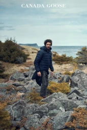 Man wearing a navy puffer jacket standing outdoors on a rocky coastal terrain under an overcast sky.