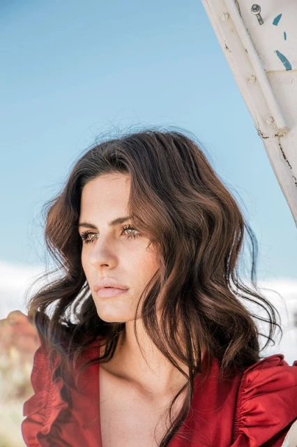 Portrait of a woman with brown wavy hair wearing a red blouse, looking away in an outdoor environment under bright daylight.