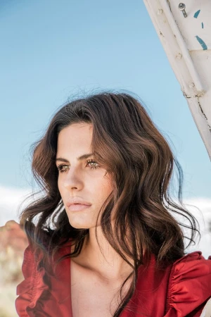 Portrait of a woman with brown wavy hair wearing a red blouse, looking away in an outdoor environment under bright daylight.