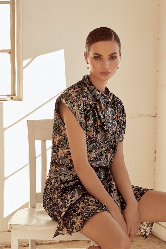 Model seated on a white wooden chair wearing a patterned short-sleeved dress in a sunlit room with pale walls.