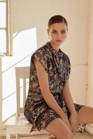 Model seated on a white wooden chair wearing a patterned short-sleeved dress in a sunlit room with pale walls.