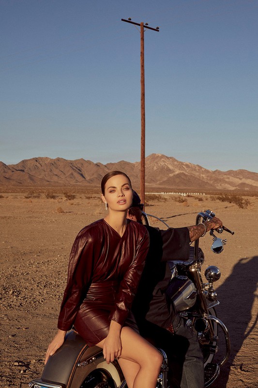 Glamorous woman in red dress sits on motorcycle in a desert landscape with mountains and utility pole under a clear blue sky.