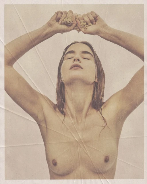 A nude young woman with wet hair raises arms and holds a clump of light-brown grains, lit in warm studio light against a beige background.