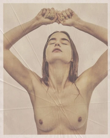 A nude young woman with wet hair raises arms and holds a clump of light-brown grains, lit in warm studio light against a beige background.