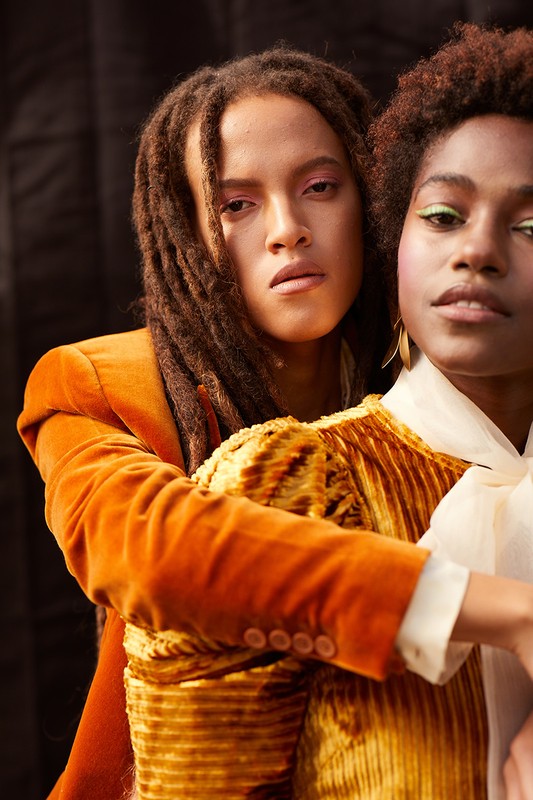 Two women pose in a studio against a black backdrop, one with dreadlocks in an orange velvet blazer and the other with an elaborate updo.