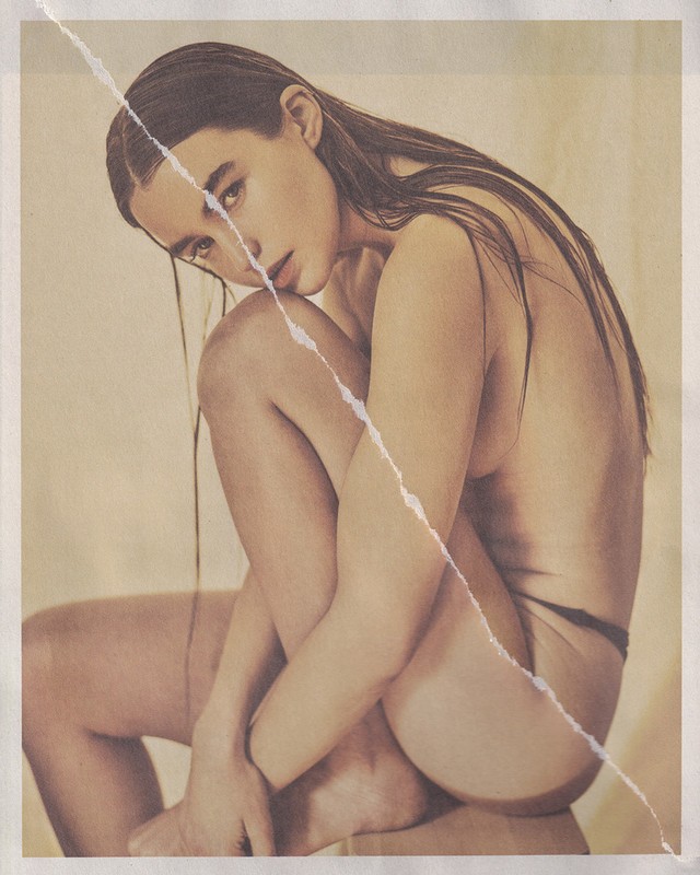 Nude woman with long brown hair sits hugging her legs in a studio, displayed on a torn beige paper background, in an artful photograph.