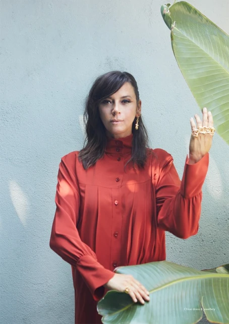 Portrait of an adult woman in a terracotta dress posing with a ring, standing next to a large plant with a textured blue background.