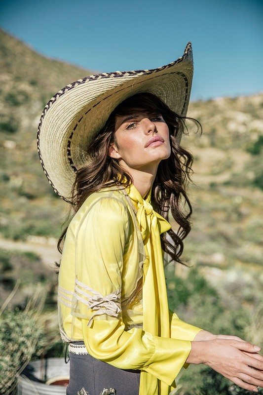 Woman poses wearing a yellow blouse and large sun hat in a desert landscape, against a blue sky, in an outdoor fashion shot.