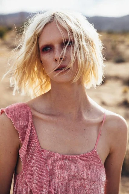 Close-up shot of a woman with fair skin, blonde wavy hair, and pink beaded top in a desert landscape with mountains in the background.