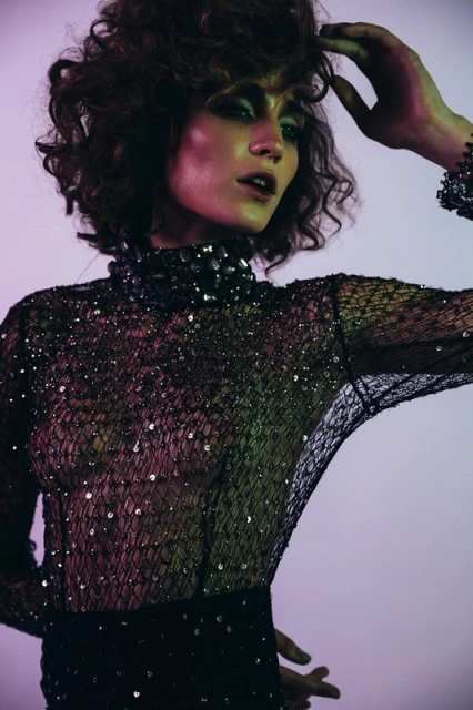 A woman with curly hair poses in a black net dress with embellishments in a studio setting with purple and green lighting, showing an editorial fashion style.