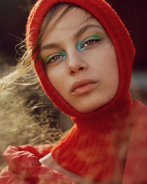 Portrait of a young woman with blue and green eyeshadow, wearing a red knitted balaclava with a blurred outdoor background.